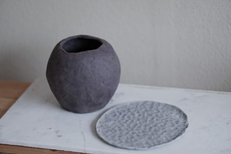Dunkelgraue, runde Vase neben einem grauen, strukturierten Dekoteller.