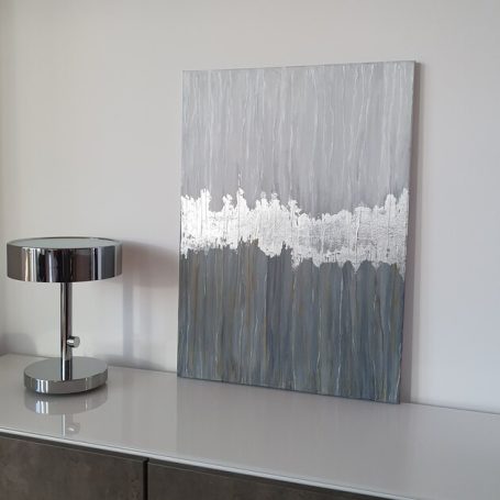 Abstrakte Wanddekoration in Grautönen mit metallischen Silberakzenten und einer Tischlampe.